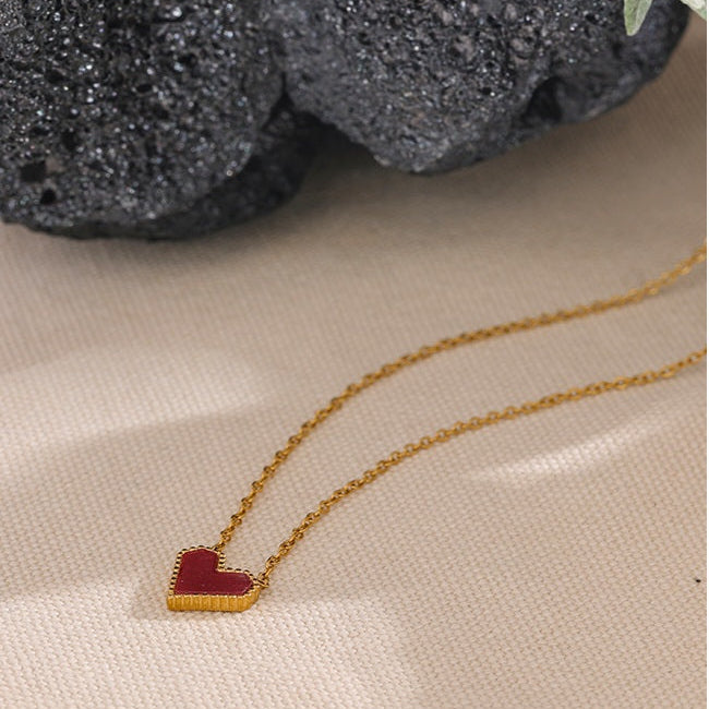 Gold necklace with a red heart pendant on a beige surface.